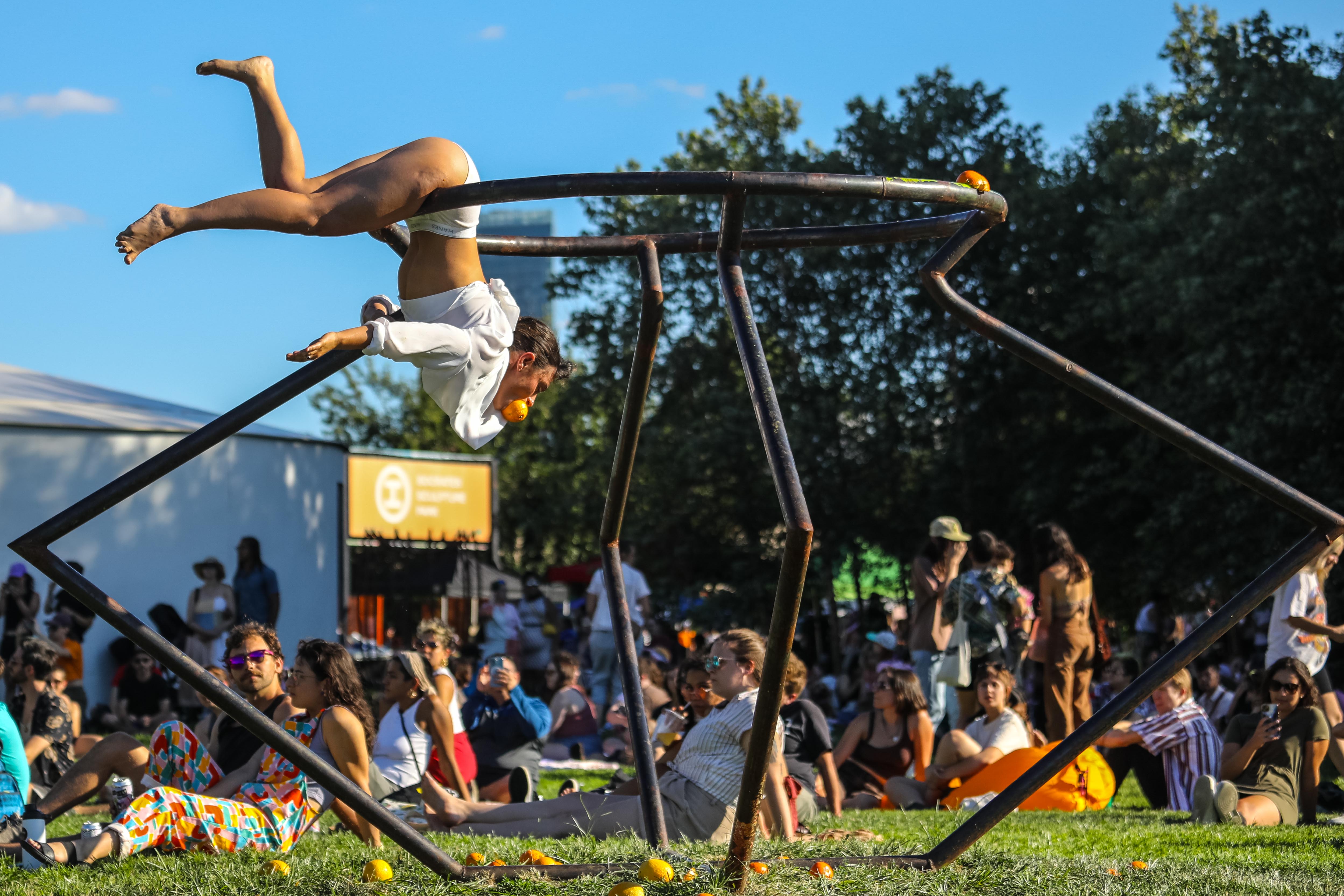 A performer acrobatically hangs from a steel sculpture while holding an orange in their mouth. A crowd of people sit on grass in the background.